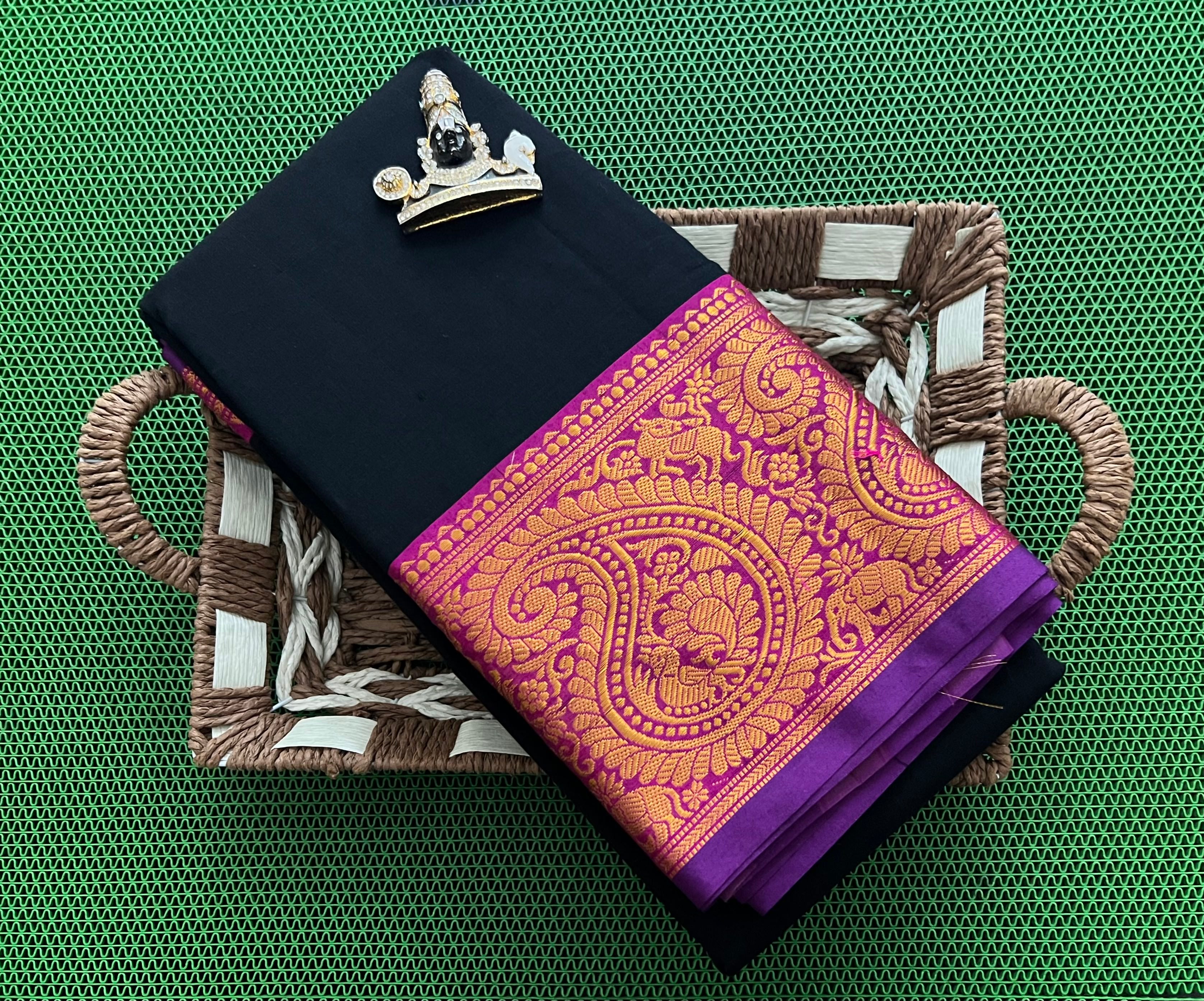 Black saree with purple and gold zari border, placed in a decorative woven tray on a green textured surface.